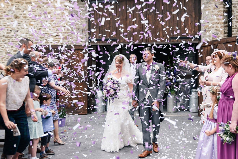 Purple and white confetti falls as the newlyweds walk outside for the first time, glowing with joy.